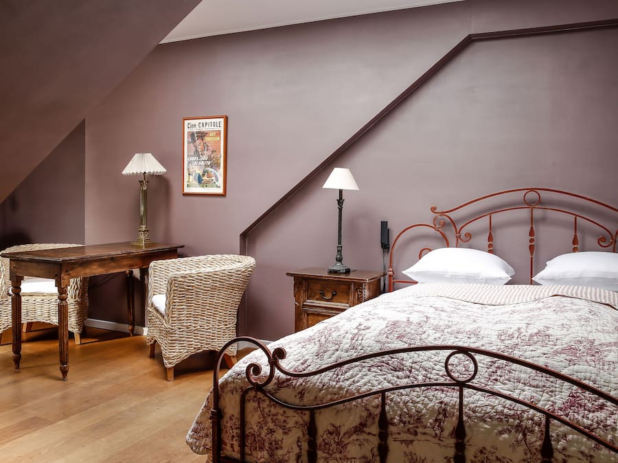 Un dormitorio con techo inclinado, un escritorio de madera con lámpara, una silla de mimbre, una cama con colcha estampada y un cartel en la pared.