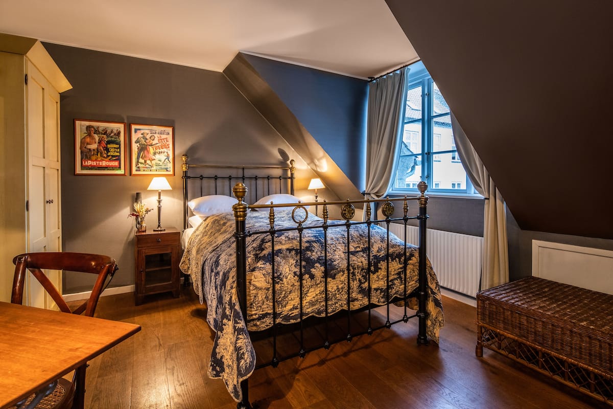 A bedroom with a sloped ceiling, a bed with a patterned bedspread, a nightstand with a lamp, a wicker bench, and two posters on the wall.