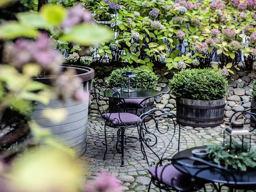Un patio al aire libre con suelo de piedra, sillas y mesas de metal. Hay plantas en macetas y una pequeña fuente.