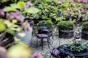 An outdoor patio with stone flooring, metal chairs, and tables. There are potted plants and a small fountain.