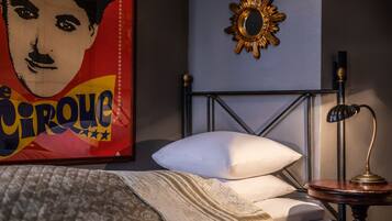 Una cama bien hecha con un almohadón blanco y una colcha estampada, un espejo con forma de sol en la pared, y una mesita de noche con una lámpara.