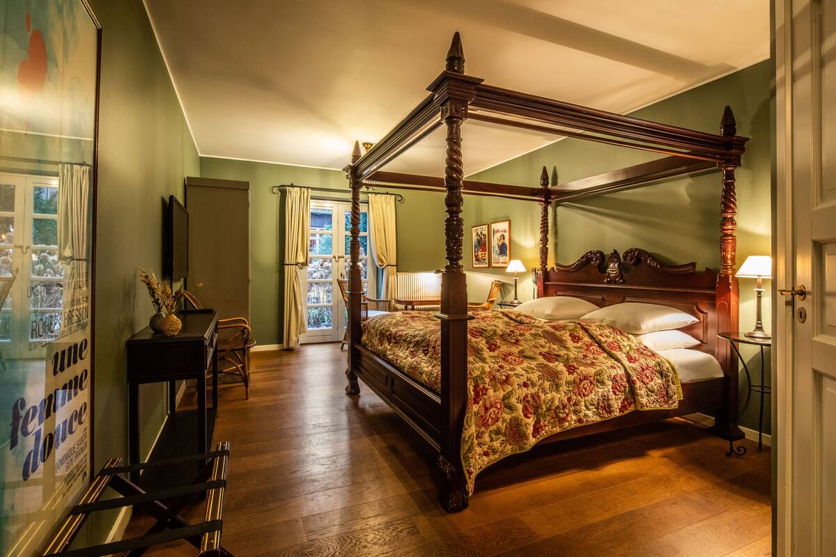 A four-poster bed with a floral bedspread, a nightstand with a lamp, and a window with curtains.