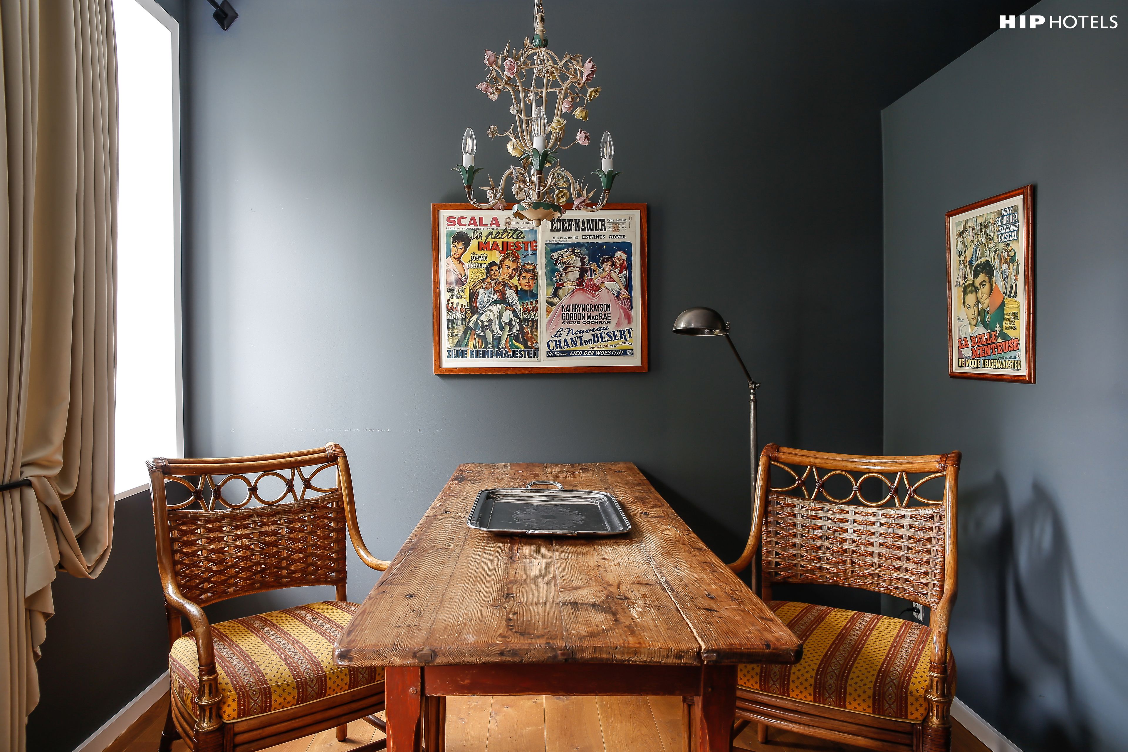 A dining area with a wooden table, two wicker chairs, a chandelier, and vintage movie posters on the wall.