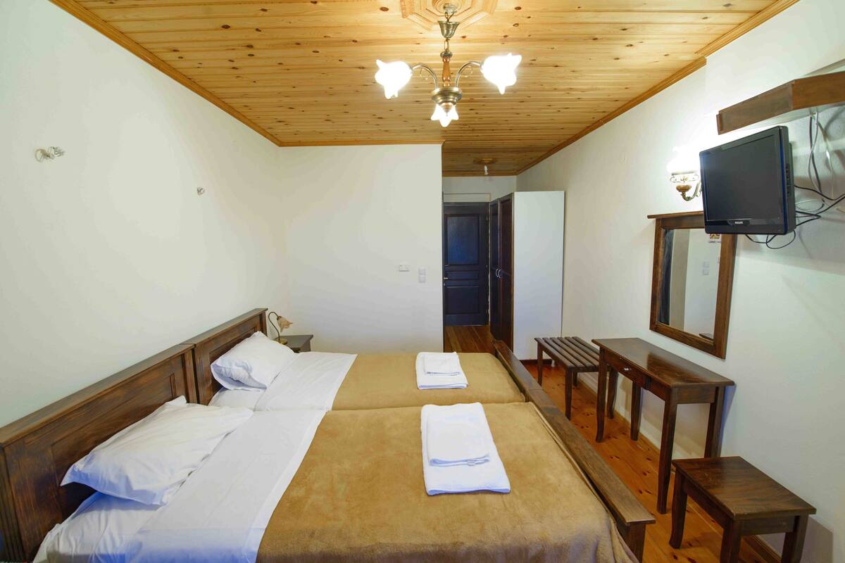 Habitación de hotel con dos camas, cabeceros de madera, ventilador de techo, televisor montado en la pared y un banco de madera.