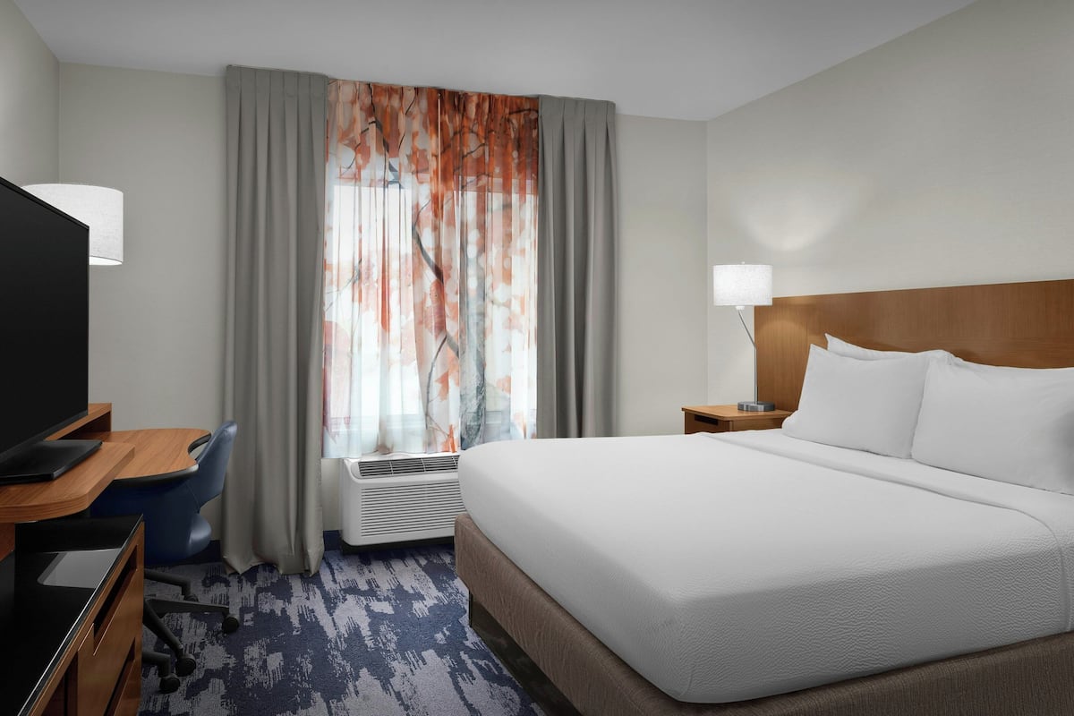 Une chambre d’hôtel comprenant un lit, un bureau avec un ordinateur, une chaise, une télévision, une lampe et une fenêtre avec des rideaux.
