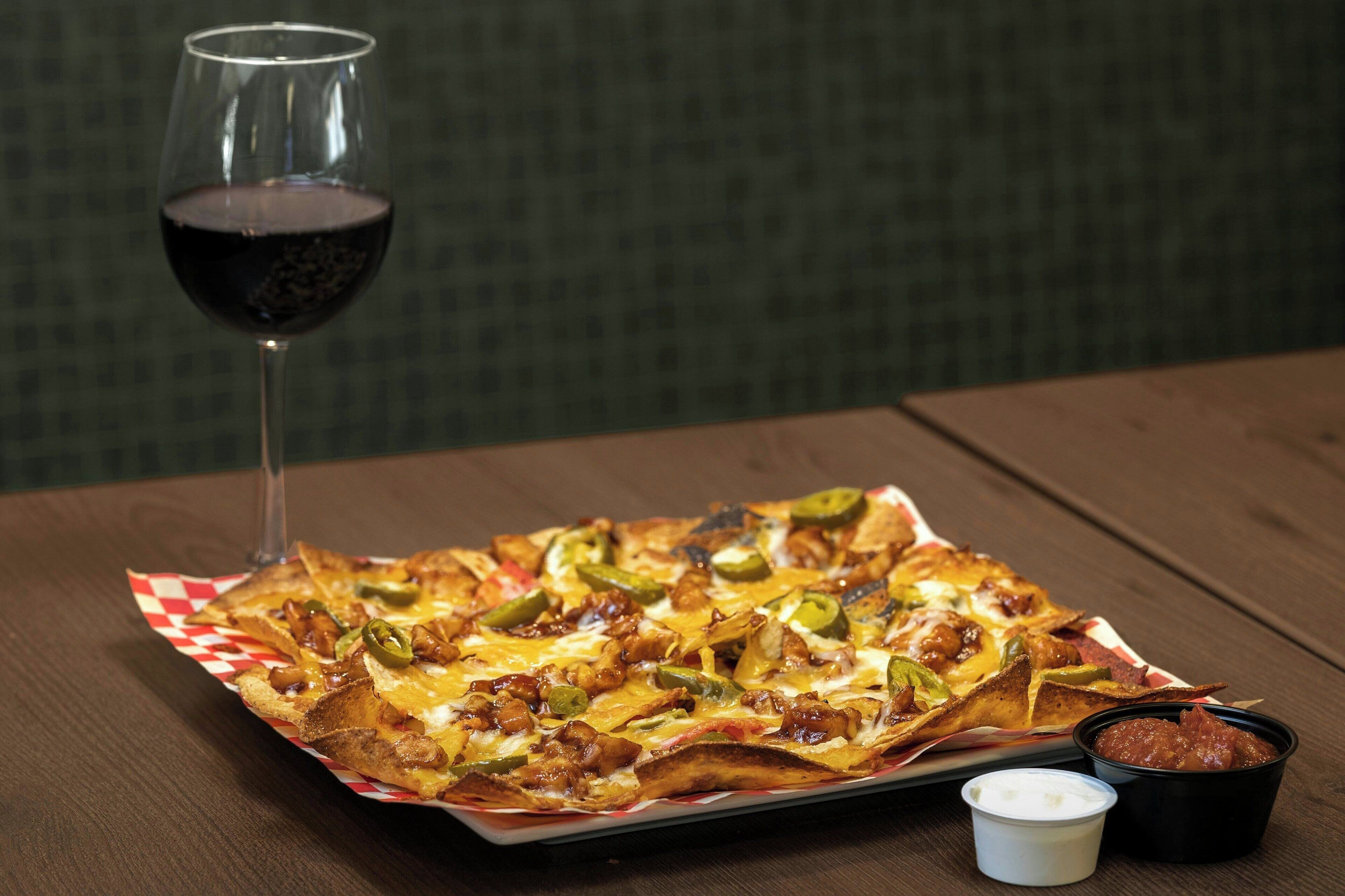 Nachos con queso, jalapeños y carne, servidos en un plato de papel con dos salsas para mojar y una copa de vino tinto.