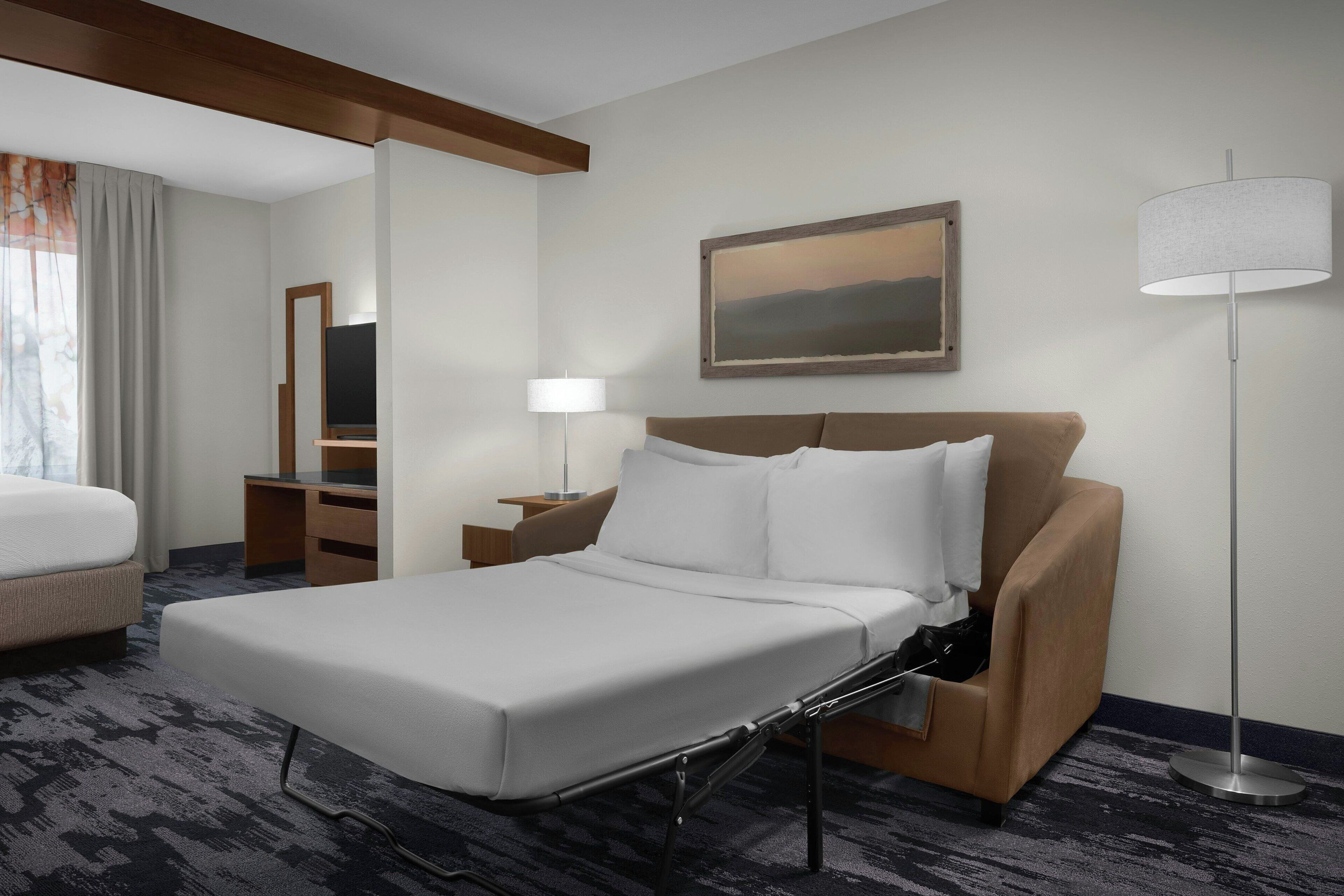 A hotel room with a bed, a desk, a chair, a lamp, and a painting on the wall.