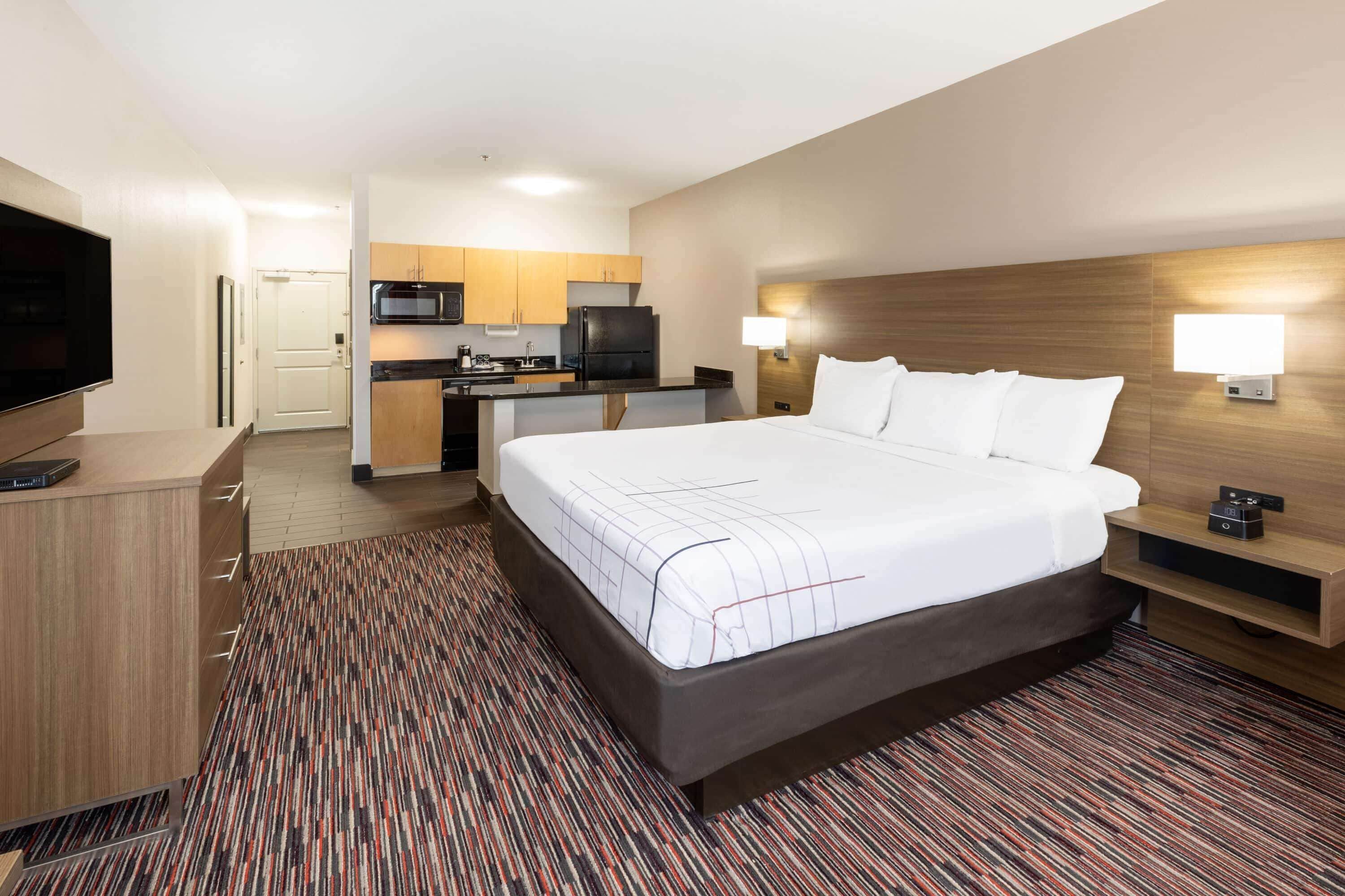 A hotel room with a large bed, a desk, a TV, a kitchenette, and a bathroom.