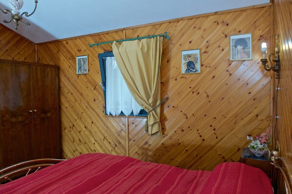 Una habitación con paredes de paneles de madera, una cama con colcha roja, una ventana con cortina y un cuadro colgado en la pared.