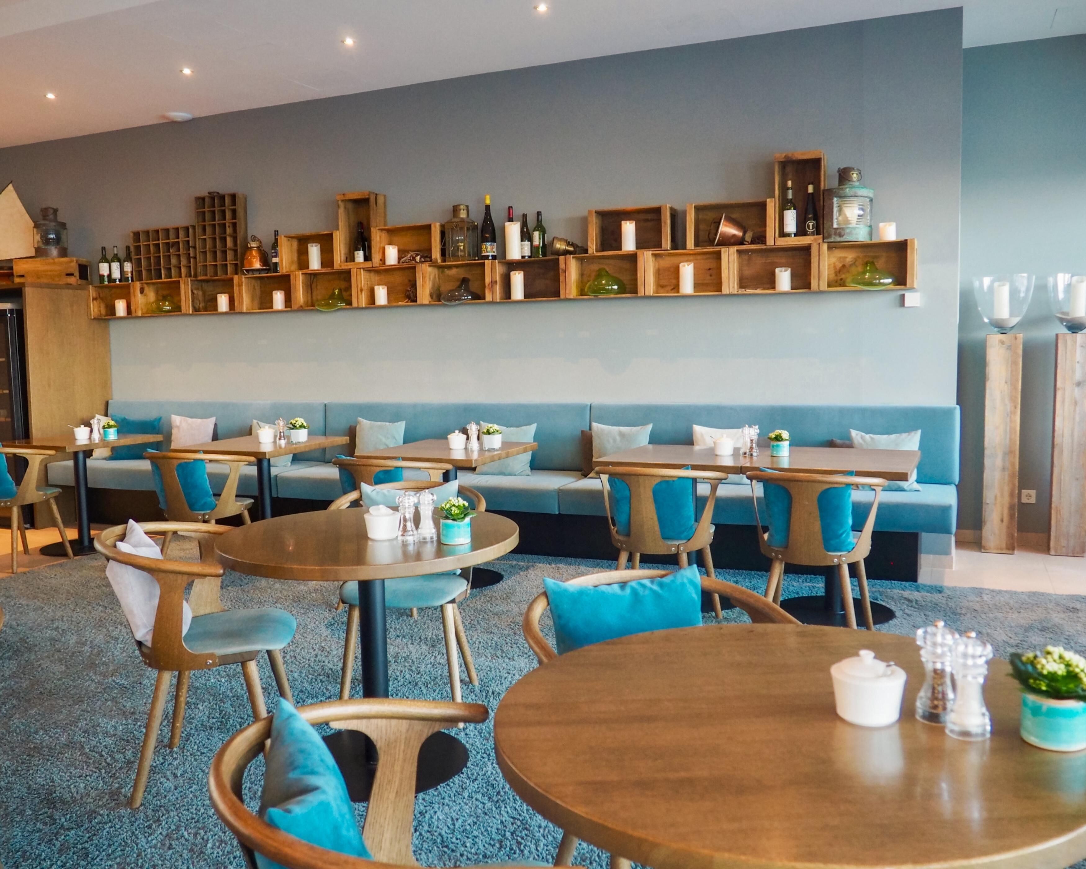 A modern lounge area with round tables and blue cushioned seating, wooden shelves with bottles and candles, and a neutral color palette.