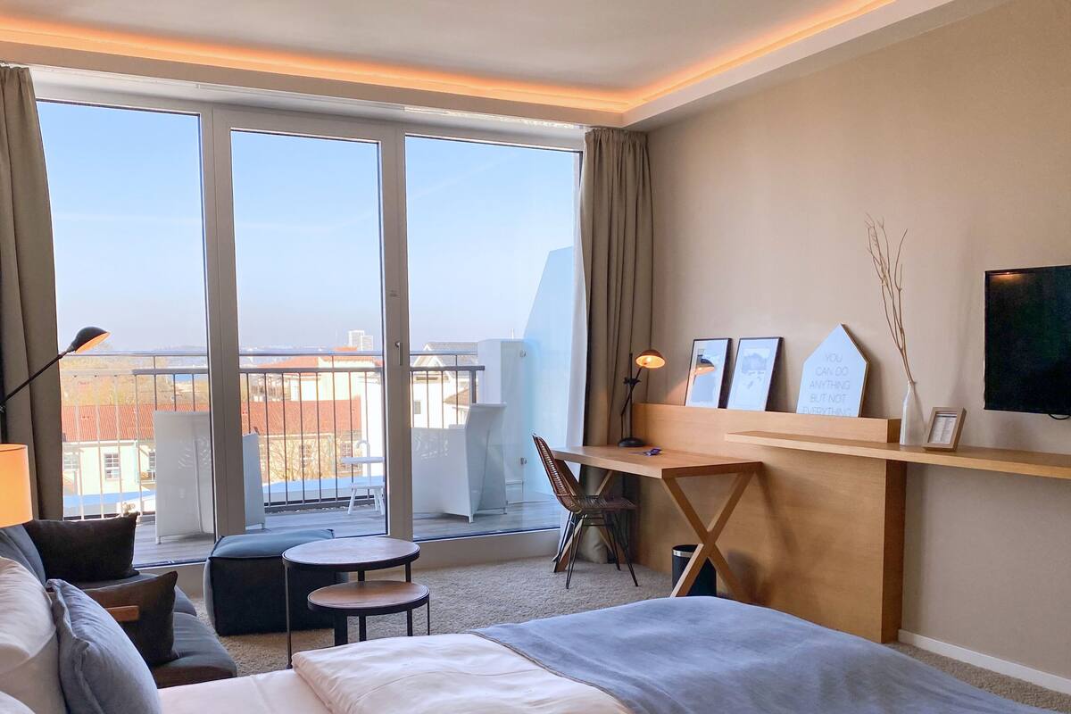 Ein Hotelzimmer mit Bett, Schreibtisch, Stuhl, kleinem Tisch und Blick auf die Stadt durch große Fenster.