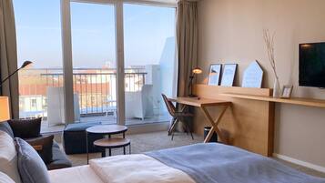 Une chambre d’hôtel comprenant un lit, un bureau, une chaise, une petite table et une vue sur la ville grâce à de grandes fenêtres.
