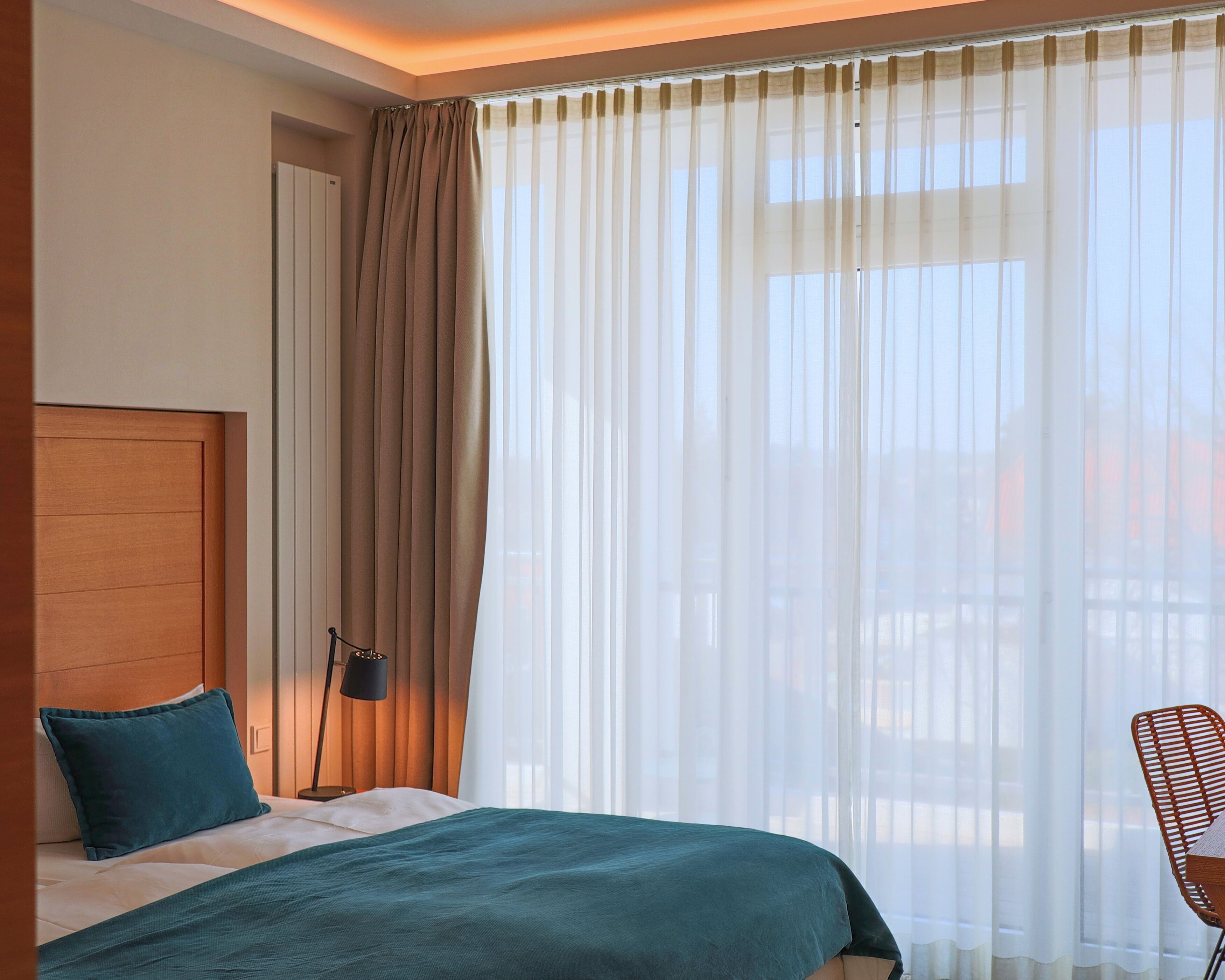 A hotel room with a bed, a nightstand, a lamp, a chair, and a large window with sheer curtains.