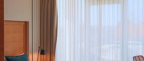 Ein Hotelzimmer mit einem Bett, einem Nachttisch, einer Lampe, einem Sessel und einem großen Fenster mit durchsichtigen Vorhängen.
