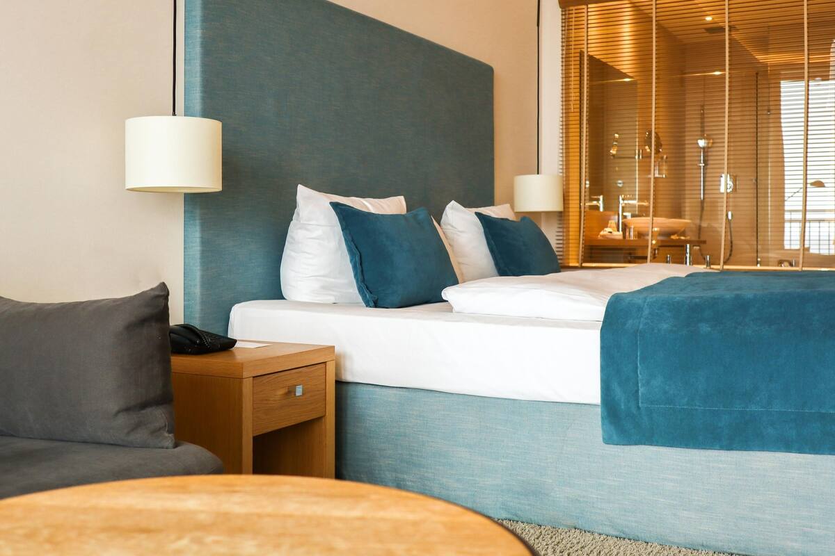 Ein modernes Hotelzimmer mit einem großen Bett, einem Nachttisch, einer Lampe und Blick auf ein Badezimmer durch eine Glastrennwand.