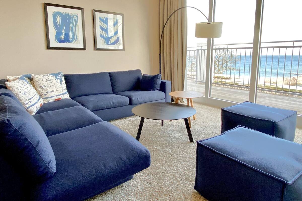 Ein modernes Wohnzimmer mit einem blauen Ecksofa, einem kleinen runden Tisch und passendem Hocker. An den Wänden befinden sich zwei gerahmte abstrakte Kunstwerke, und ein großes Fenster bietet Blick auf den Strand.