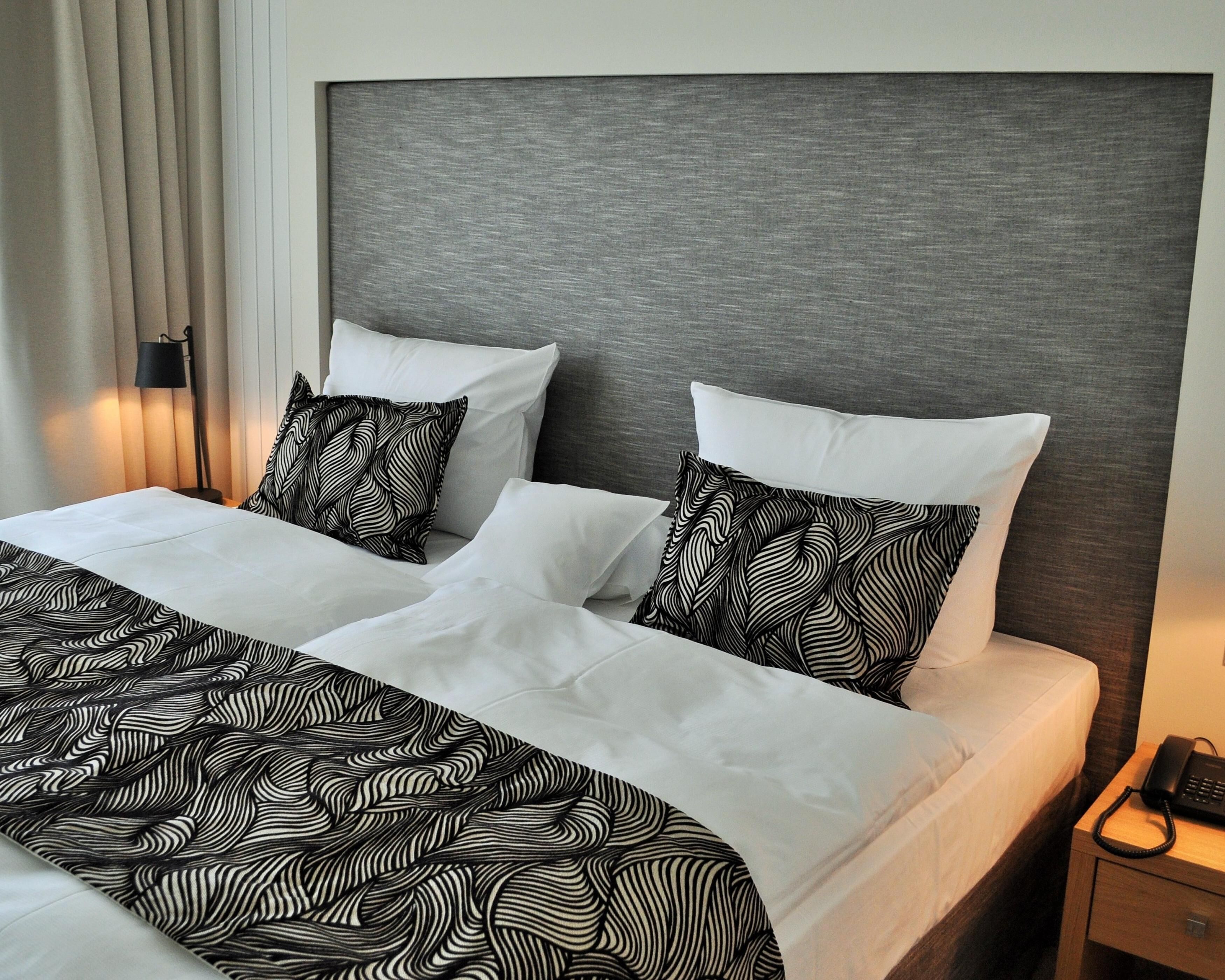 A neatly made bed with white linens and black patterned pillows, a grey headboard, and a bedside table with a phone.
