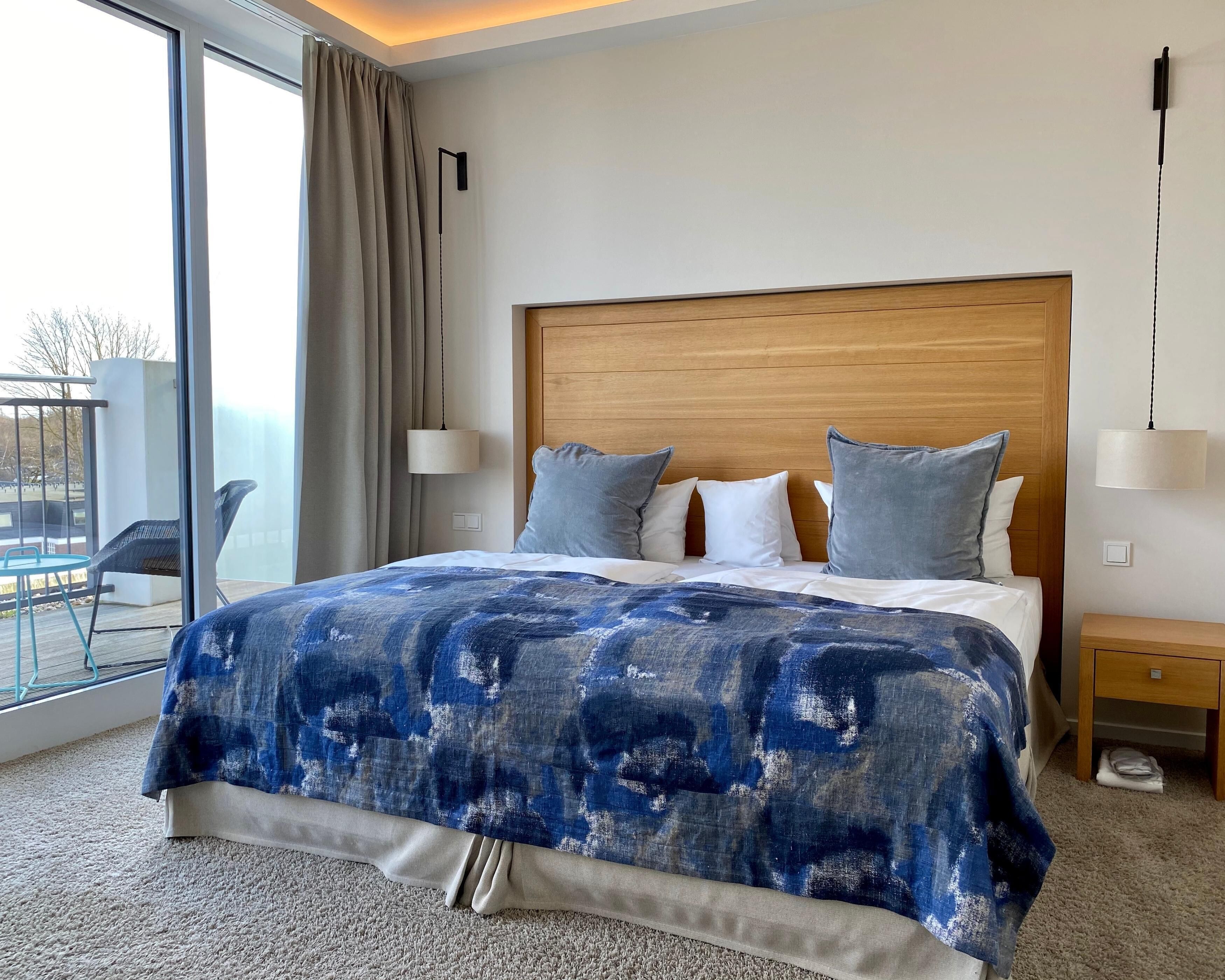 A hotel room with a large bed, a wooden headboard, and a view of the outdoors through a large window.