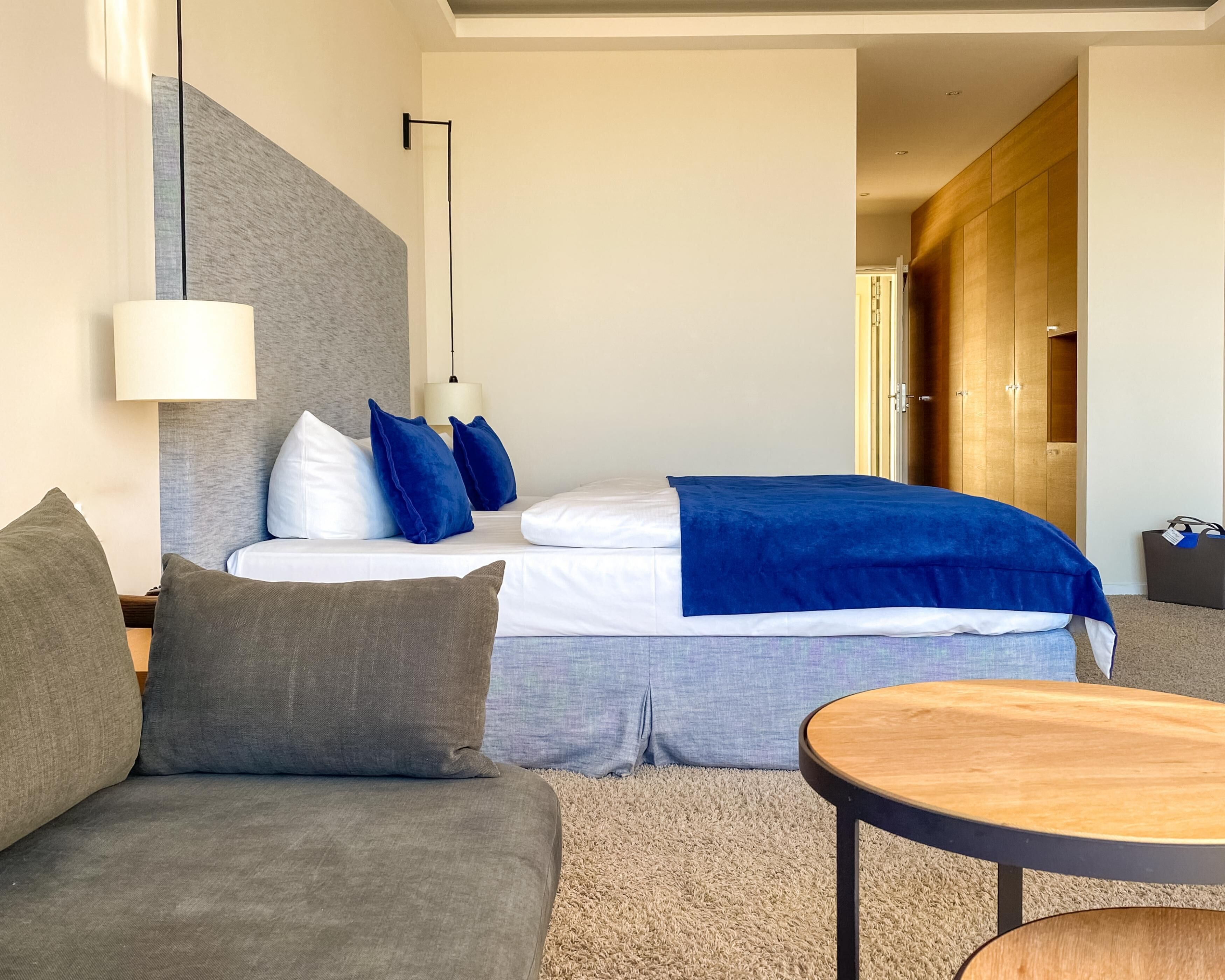 A modern hotel room with a bed, a sofa, a round wooden table, and a wardrobe.