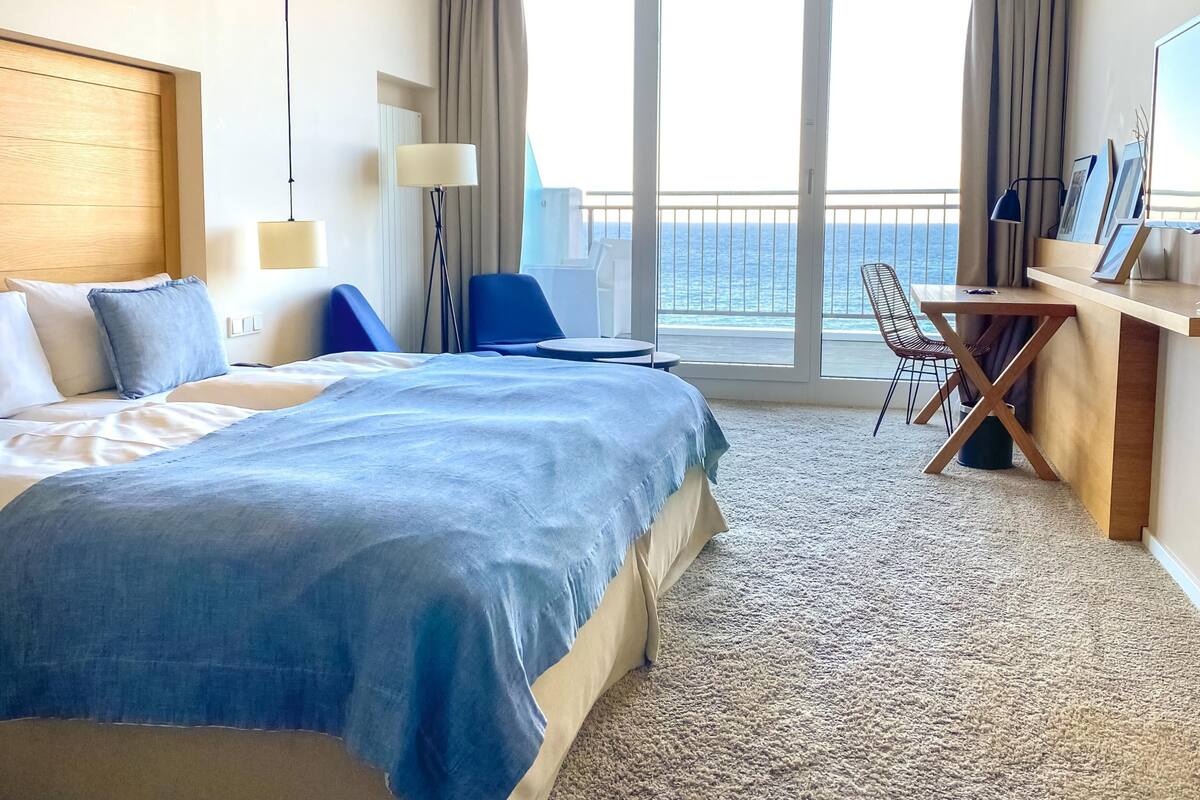 Ein Hotelzimmer mit einem großen Bett, einem Schreibtisch mit Stuhl, Blick auf den Ozean und einem Balkon mit Vorhängen.