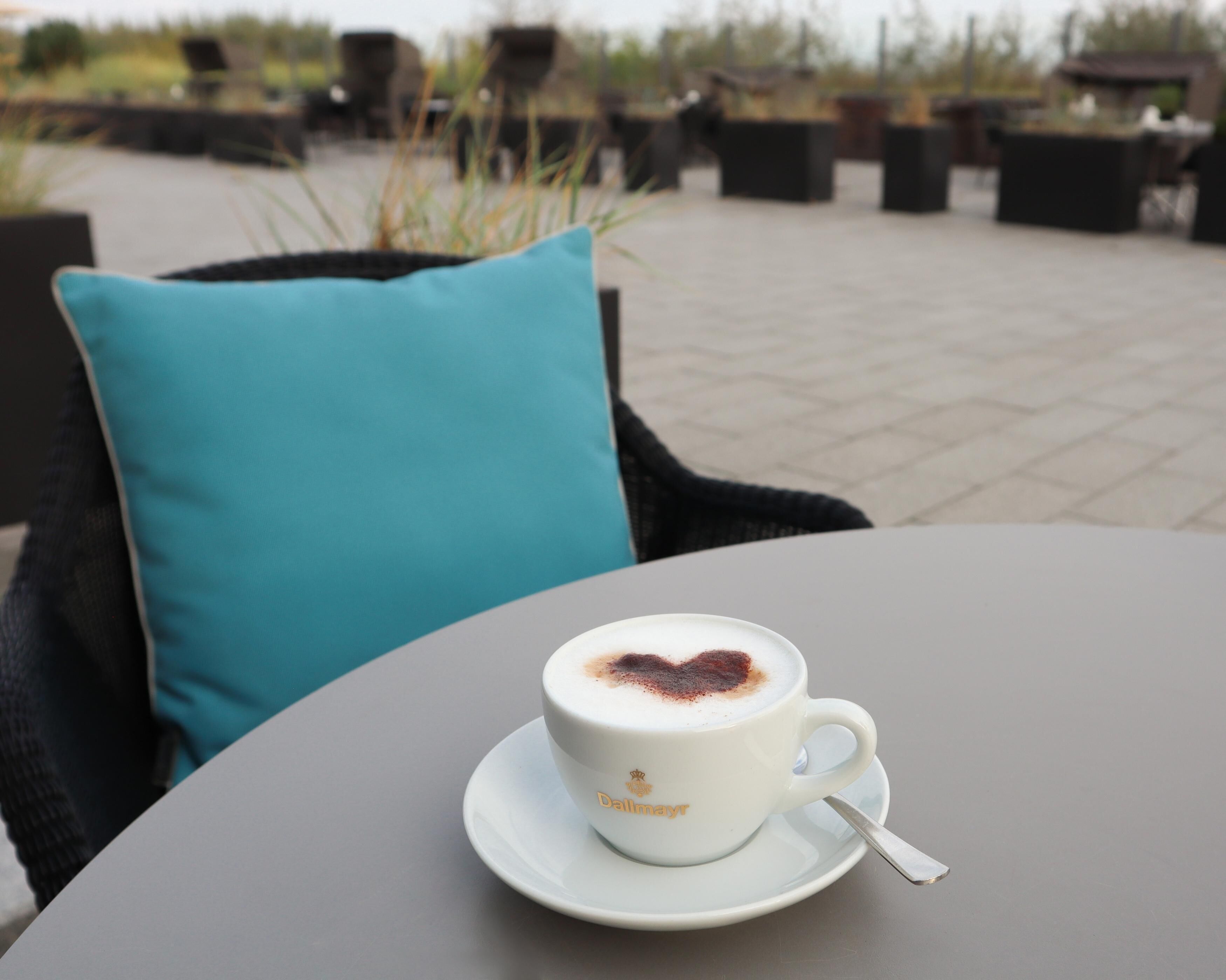 A white cup with a heart-shaped design on the foam, labeled 'Dallmayr', on a white saucer with a spoon, placed on a round table.
