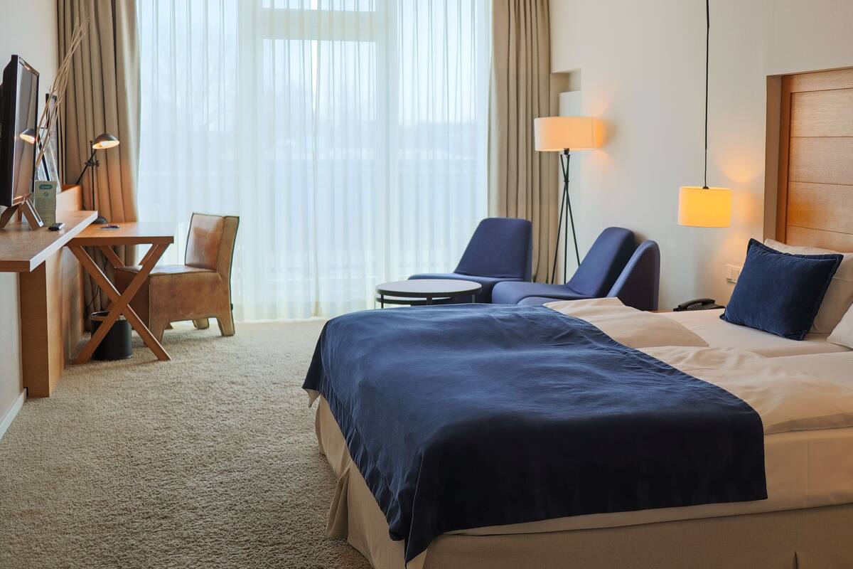 Ein Hotelzimmer mit einem großen Bett, zwei Sesseln, einem Schreibtisch mit einem Computer und einem Fernseher.
