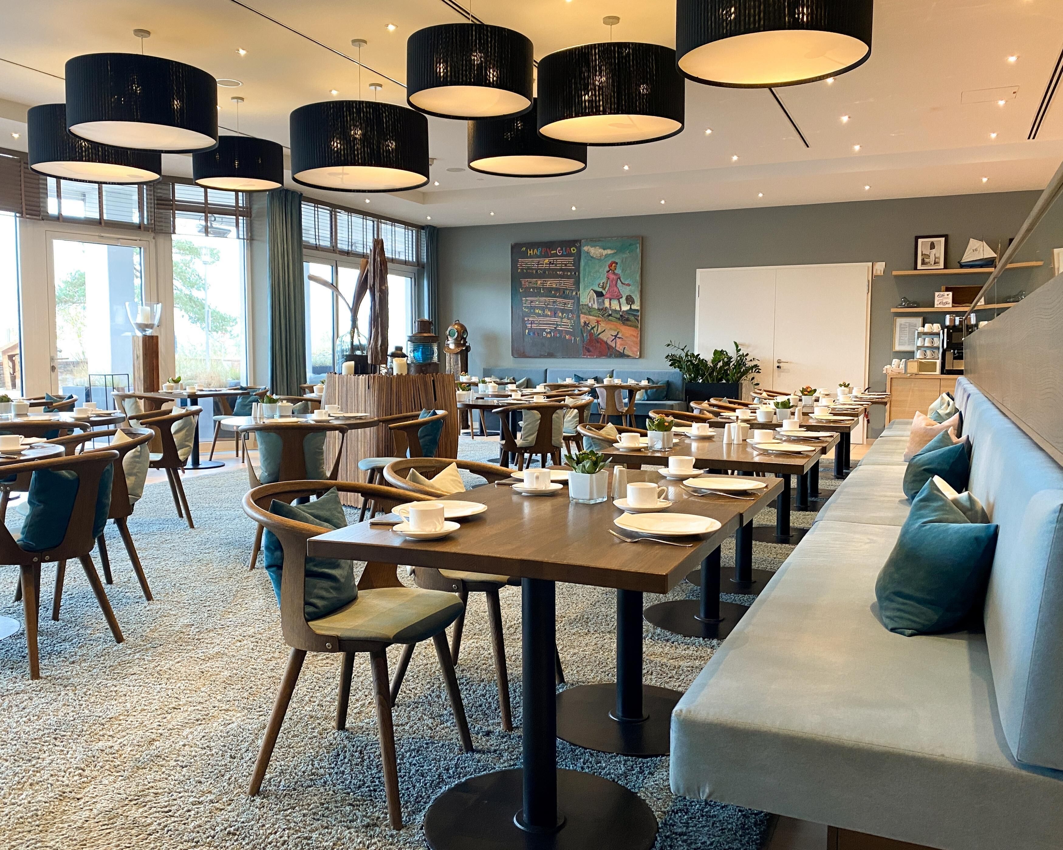 A modern dining area with multiple tables and chairs, large black pendant lights, and a view of the outdoors through large windows.