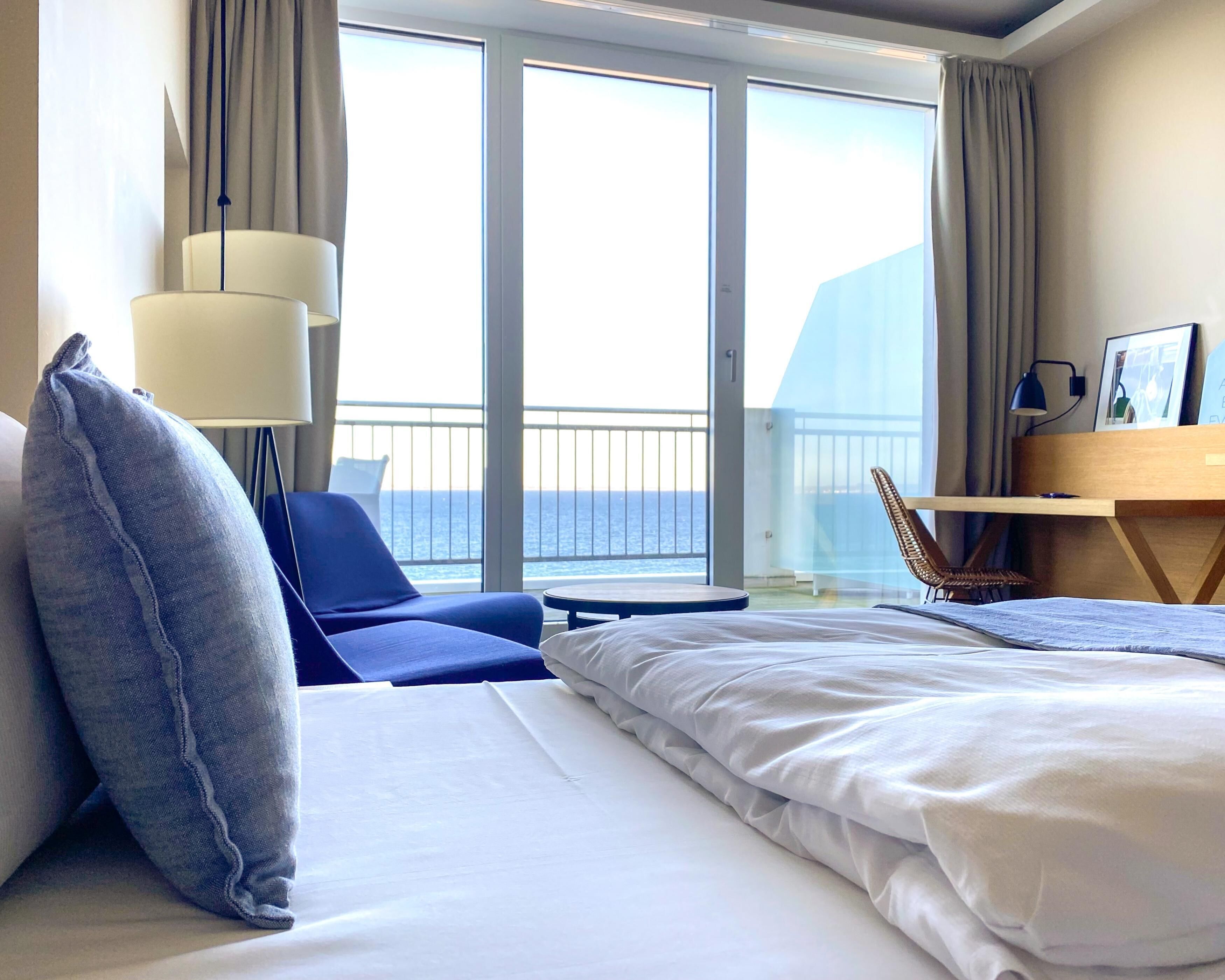 Habitación de hotel con cama, escritorio, silla y vista al mar a través de amplias ventanas.