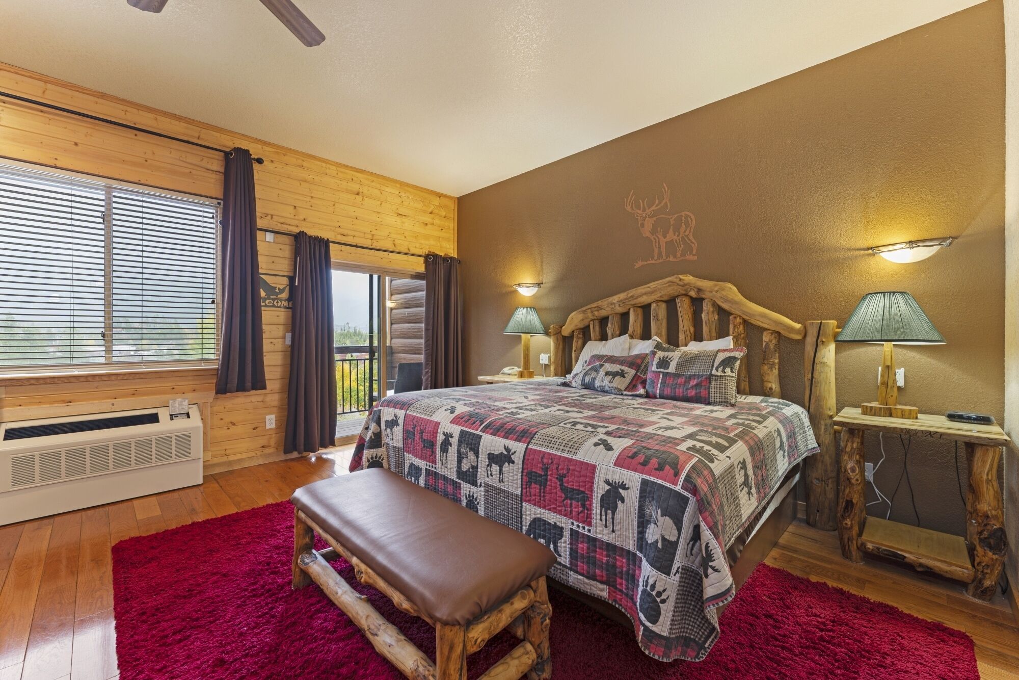 Une pièce chaleureuse avec une tête de lit en bois, un grand lit recouvert d’une courtepointe à motifs, un banc et un tapis rouge.