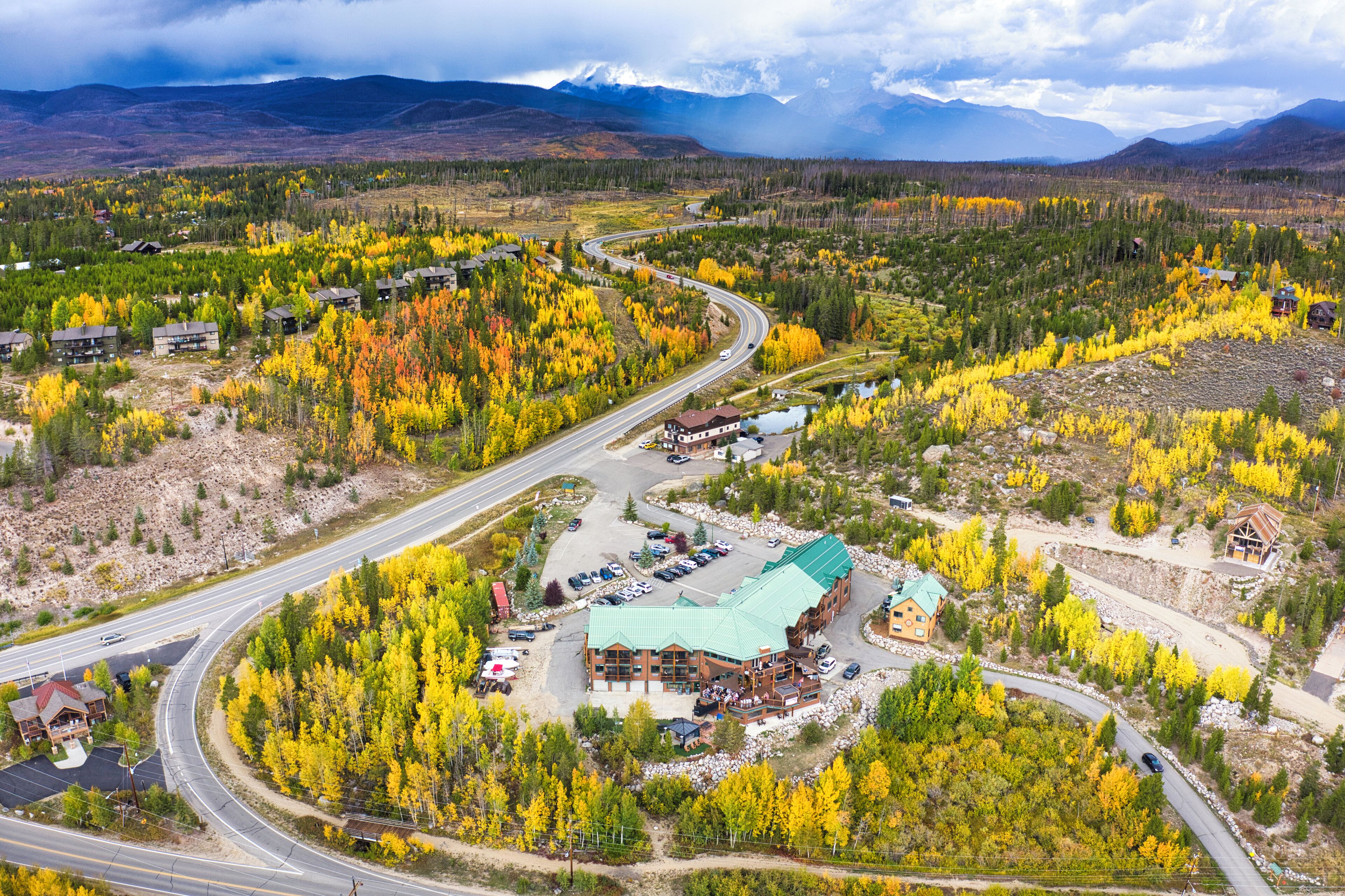 A lodge with a green roof, surrounded by autumn-colored trees, near a winding road and a mountainous backdrop.