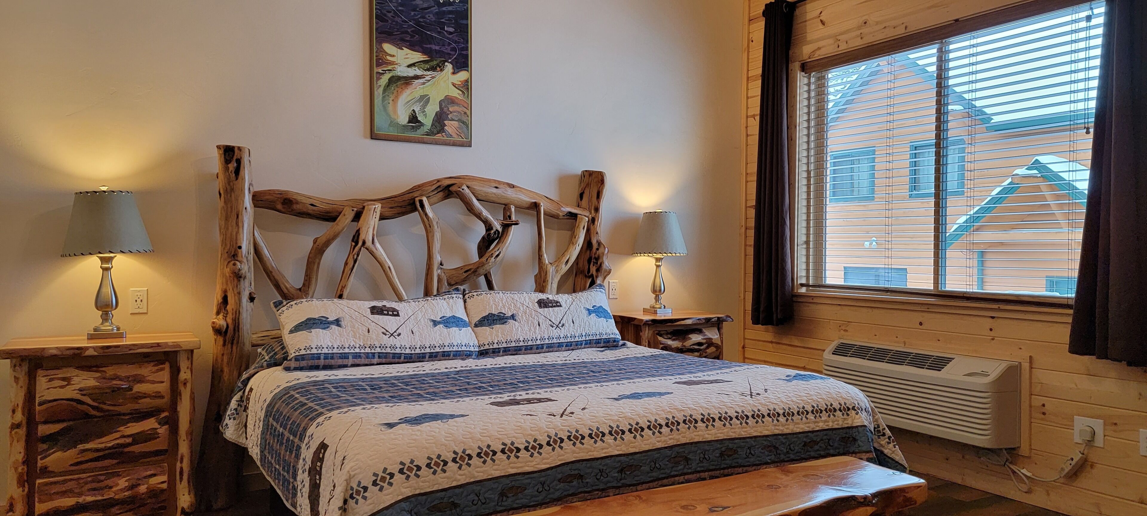 Una cama con estructura de madera y colcha estampada, dos lámparas de noche, un cuadro colgado en la pared y una ventana con persianas.