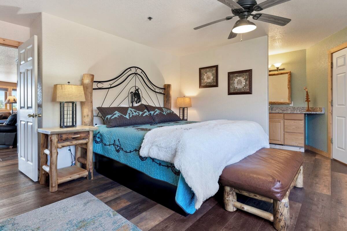 A bedroom with a bed, bedside lamps, a wooden bench, a ceiling fan, and a bathroom visible in the background.