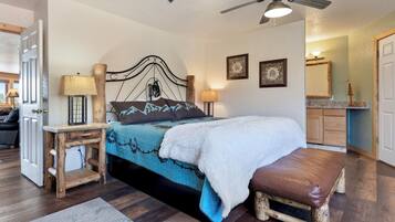 A bedroom with a bed, bedside lamps, a wooden bench, a ceiling fan, and a bathroom visible in the background.
