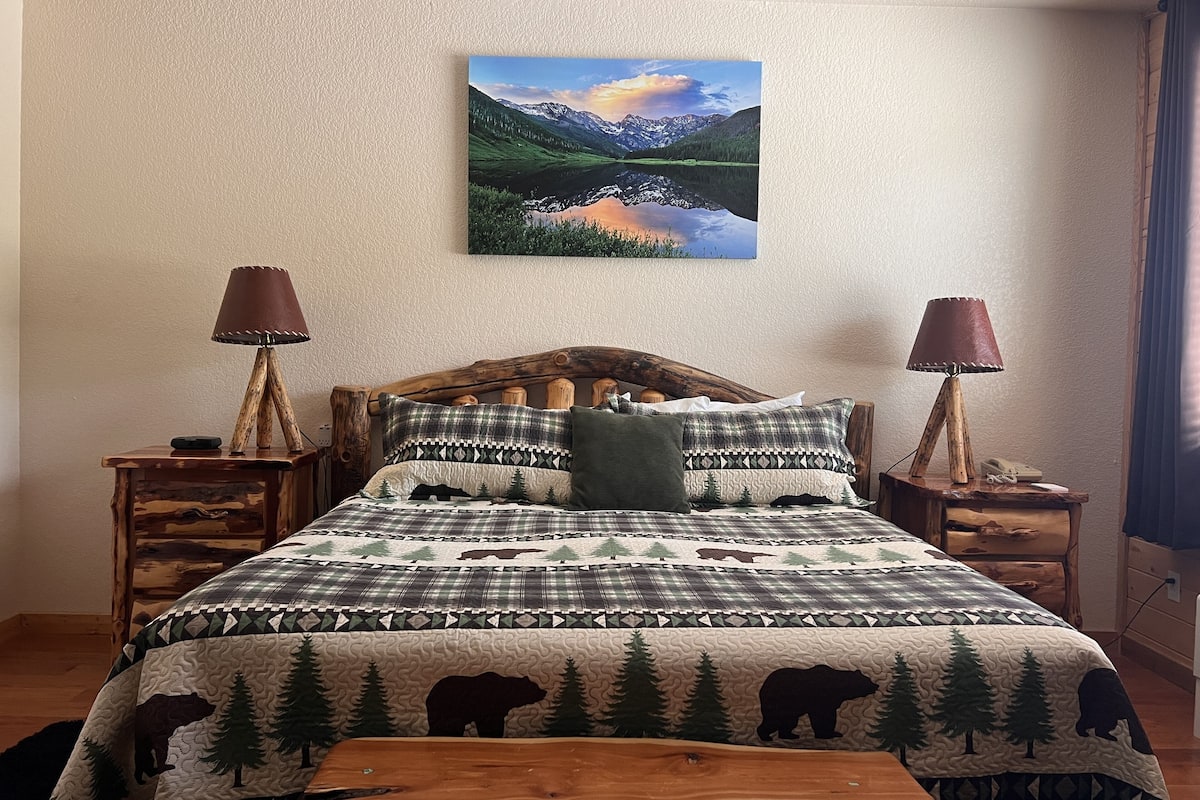 A bedroom with a wooden headboard, a mountain landscape painting, two bedside lamps, and a bed with a forest-themed quilt.
