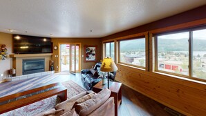 A cozy living room with a fireplace, a large TV, and a view of a town through large windows.