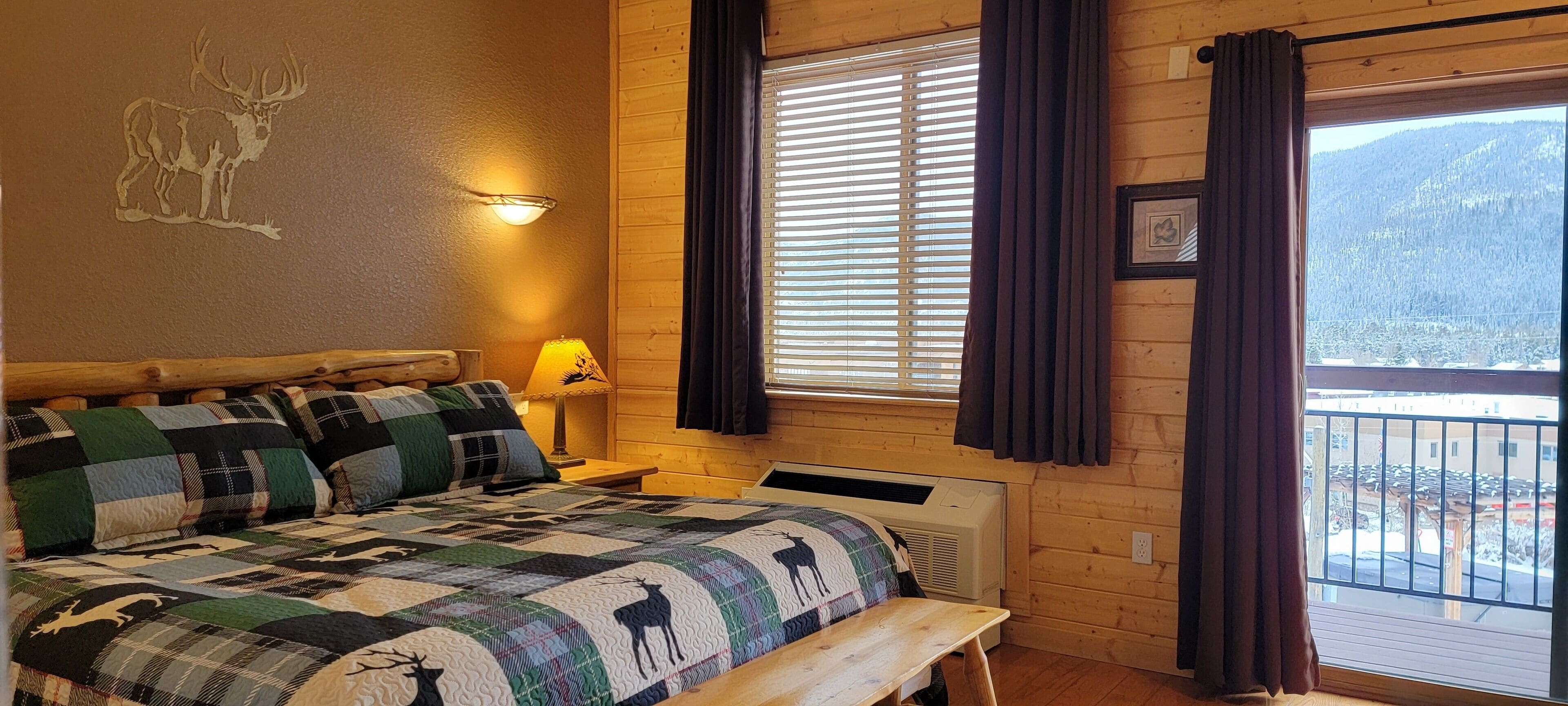 A cozy room with a bed, a quilt with animal prints, a wall-mounted lamp, a window with curtains, and a view of a snowy landscape.