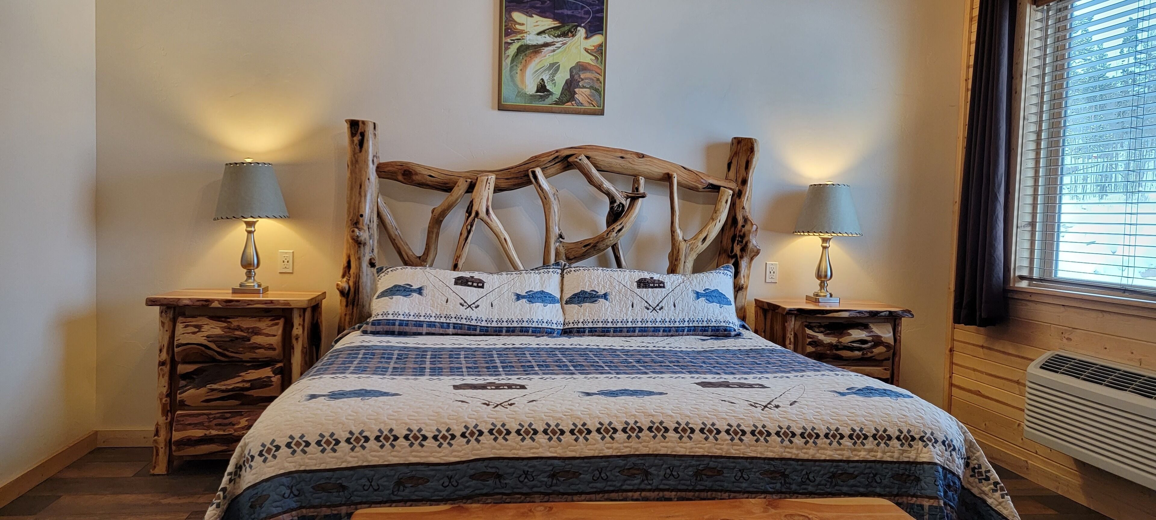 A bed with a unique wooden headboard, two bedside tables with lamps, a quilt with a fish pattern, and a painting on the wall.