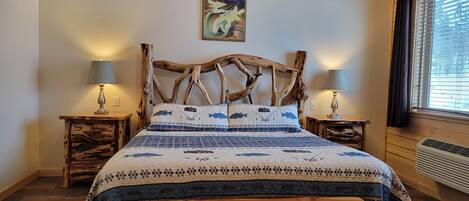 A bed with a unique wooden headboard, two bedside tables with lamps, a quilt with a fish pattern, and a painting on the wall.
