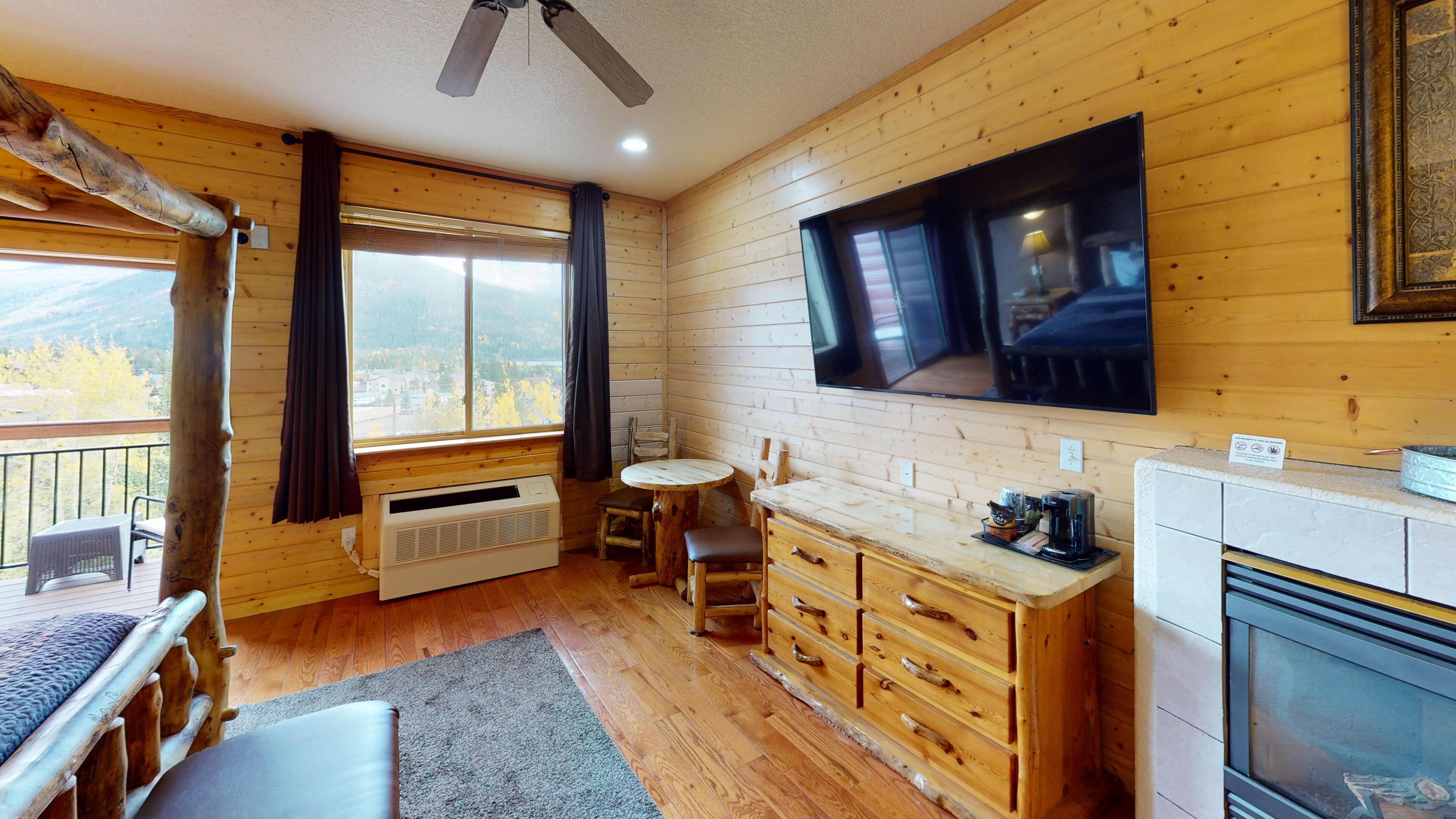 A cozy cabin interior with a wooden interior, a large window offering a view of the outdoors, a flat-screen TV mounted on the wall, and a fireplace.