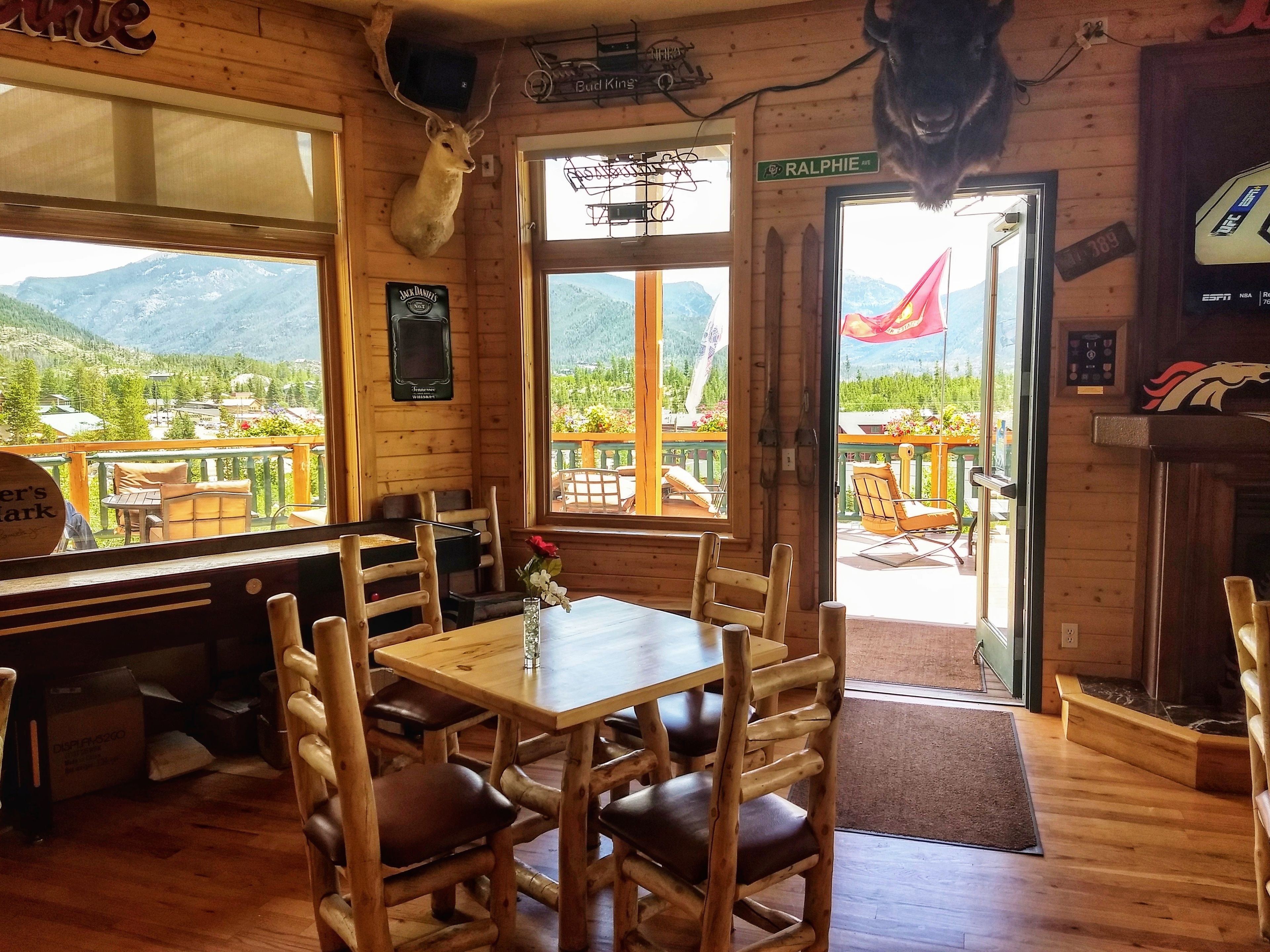 Interior de madera con una mesa, sillas, cabeza de venado montada y vistas a montañas a través de la puerta.