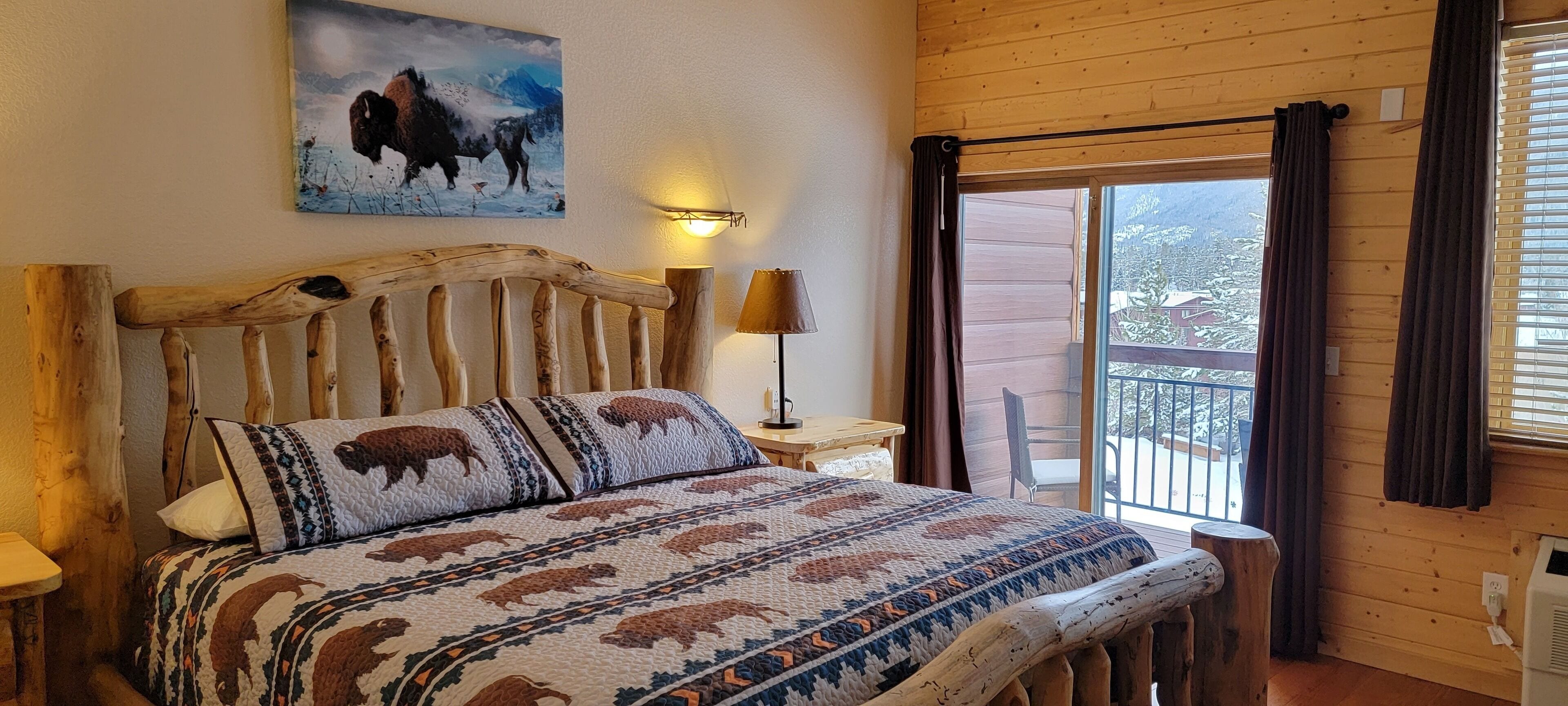 Uma cama com estrutura de madeira e roupa de cama com tema de búfalo, uma pintura de bisão na parede e vista do exterior pela janela.