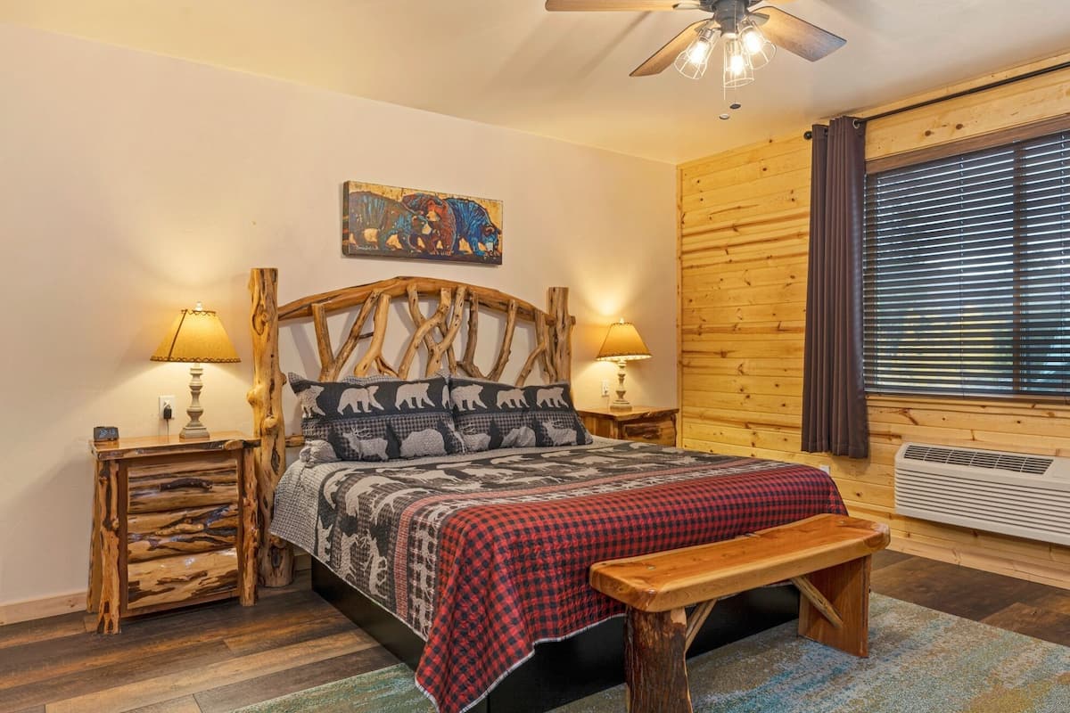 A cozy bedroom with a wooden headboard, two bedside tables with lamps, a bench, a bed with a red and black checkered blanket, a painting on the wall, and a window with blinds.