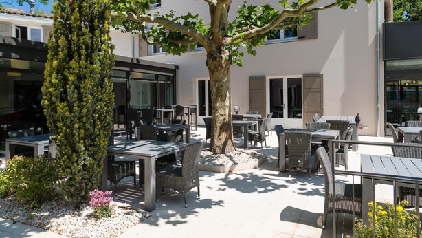 An outdoor patio area with tables and chairs, surrounded by a large tree and a building with windows. - Le Cottage de Clairefontaine (Chonas-l'Amballan)