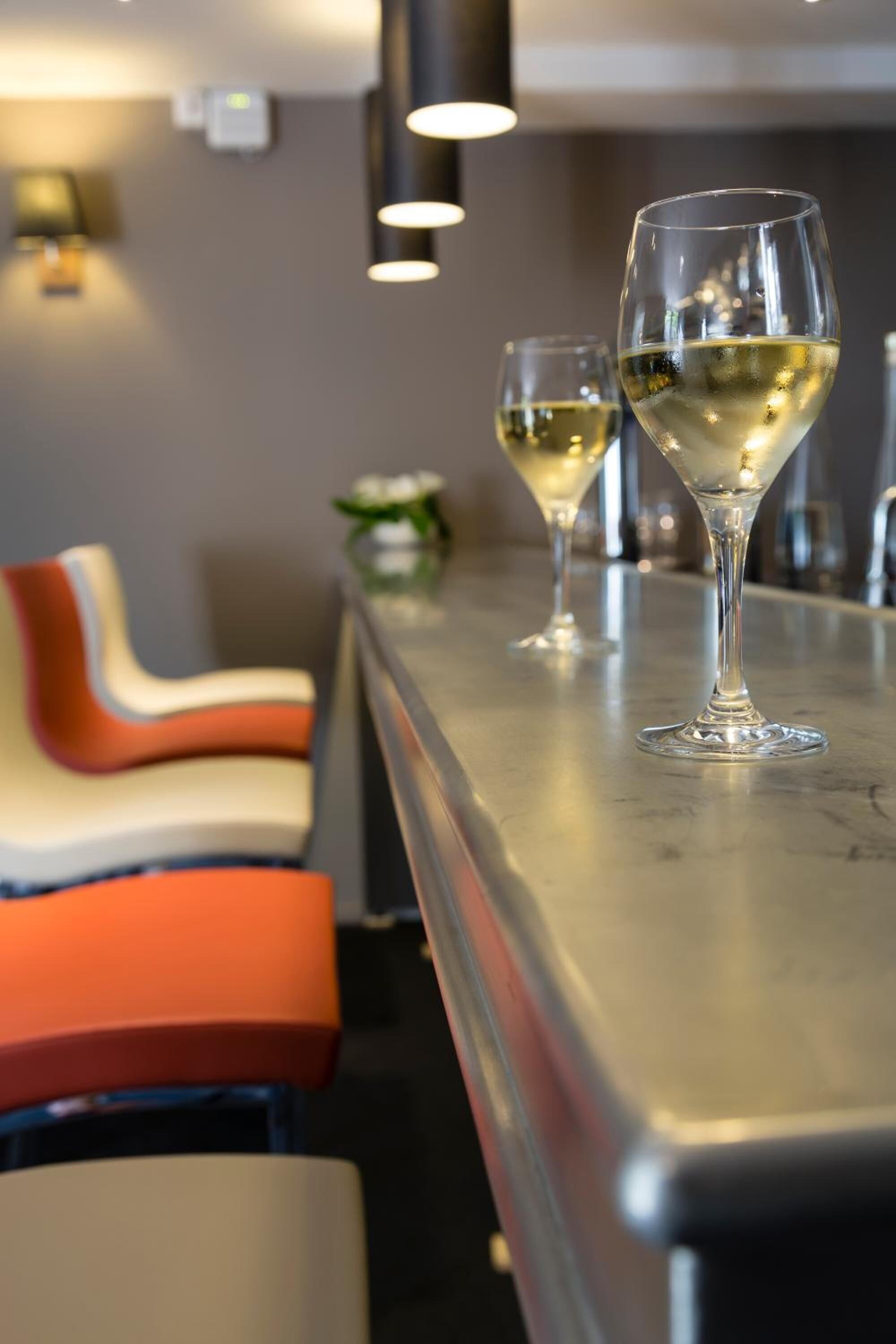 A bar area with two glasses of white wine on a counter, a modern chair design, and a wall-mounted light fixture.