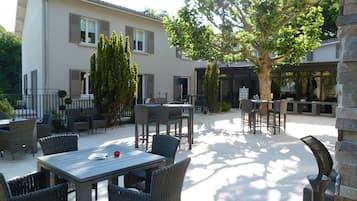 An outdoor patio area with wicker furniture and a table, a large tree, and a building with windows and shutters.