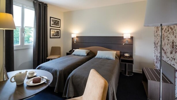 Habitación de hotel con dos camas, una mesita pequeña con una tetera y un plato de comida, una silla y una mesita de noche con una lámpara.
