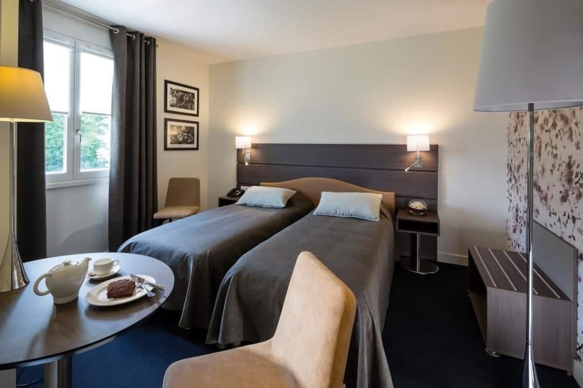 A hotel room with two beds, a small table with a teapot and a plate of food, a chair, and a bedside table with a lamp.