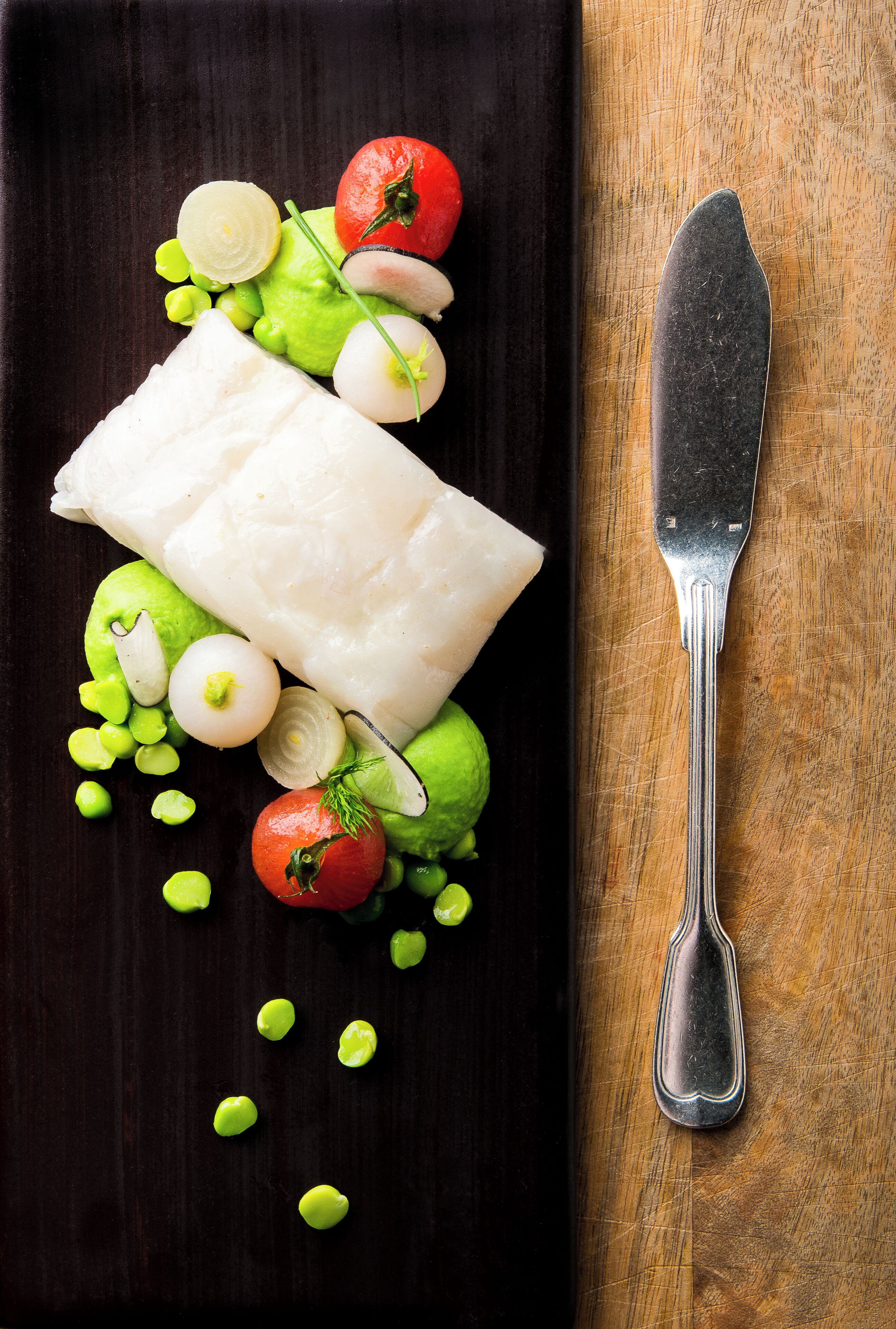 A white fish fillet on a dark plate with green peas, tomatoes, and a knife on a wooden surface.