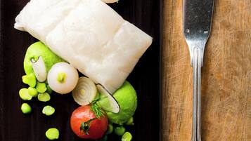 A white fish fillet on a dark plate with green peas, tomatoes, and a knife on a wooden surface.