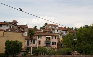 Una fila di edifici con tetti in terracotta e balconi, alcuni con parabole, e vegetazione rigogliosa in primo piano.